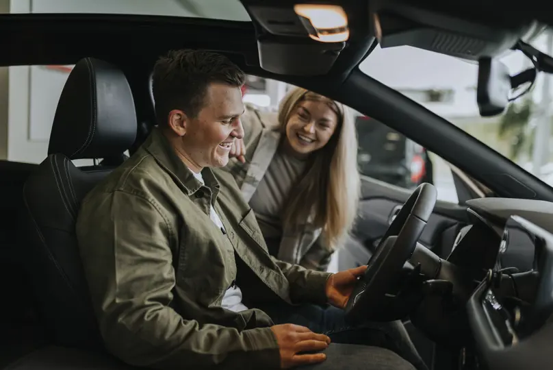 En kunde sitter i førersetet på en hvit bil og mottar bilnøklene fra en selger utenfor bilen.